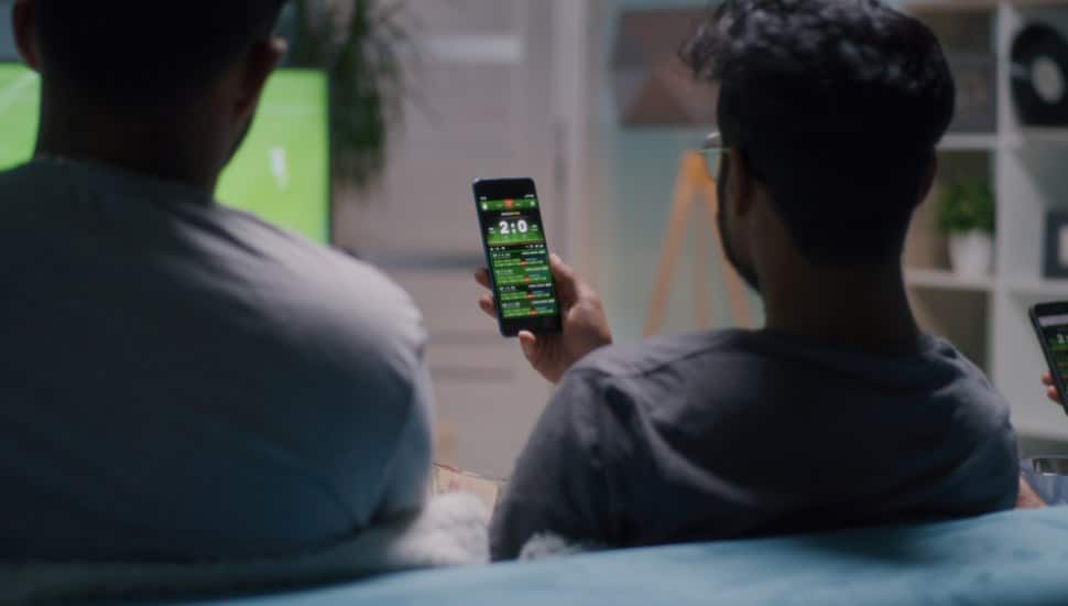 Men placing stakes on smartphones and speaking while sitting on sofa and watching soccer match together.