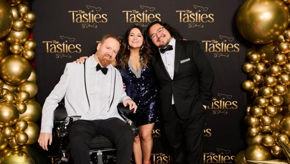 Eli Kulp, Marisa Magnatta and Dave Wez with "The Tasties" backdrop behind them with gold balloons.