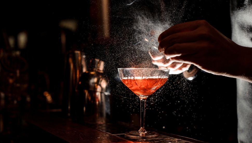 Barmans hands sprinkling the juice into the cocktail glass filled with alcoholic drink on the dark background