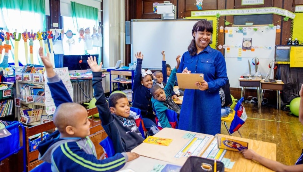 A teacher in a classroom with students raising their hands.
