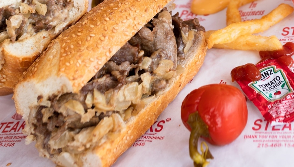 A cheesesteak on a seeded roll, surrounded with ketchup packets and a pepper.