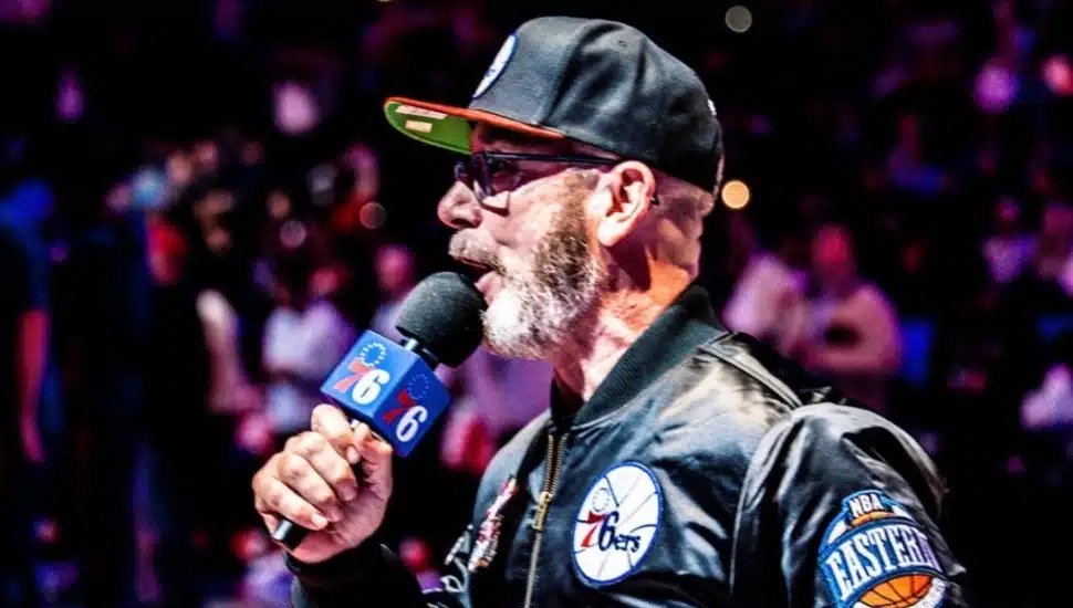 Matt Cord announcing, with mic in hand, and a Sixers hat and jacket.