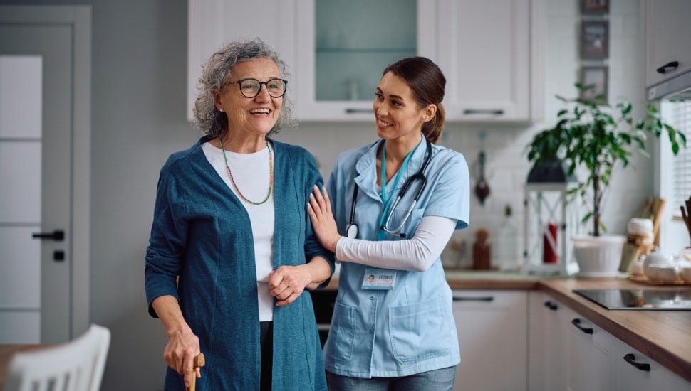 Happy home caregiver assisting her senior patient with walking through the kitchen.