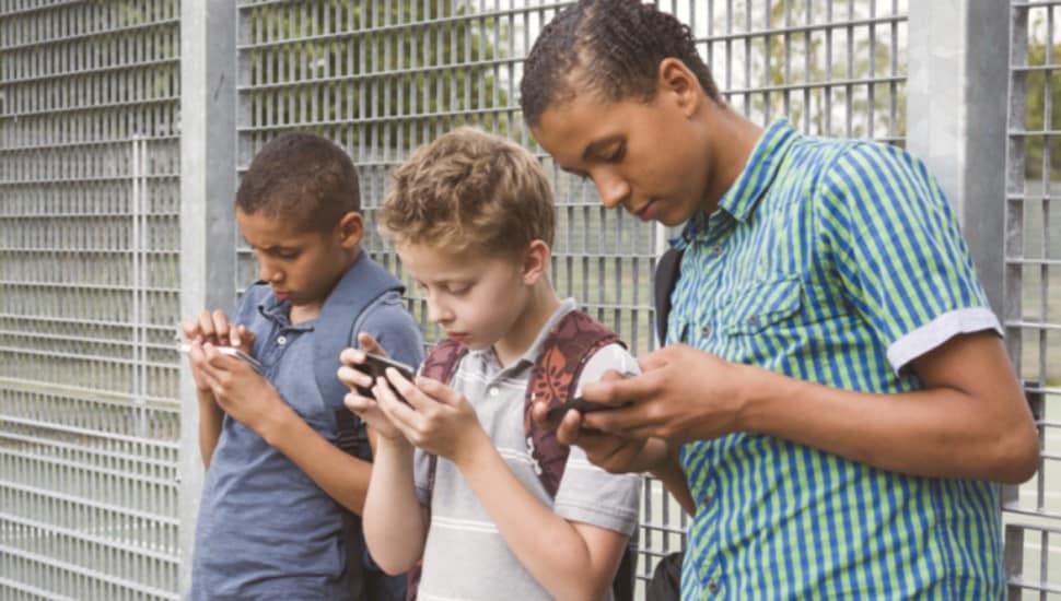 Three boys are standing along a fence looking at their phones.