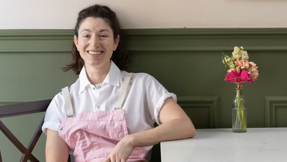 Philadelphia chef Amanda Shulman smiling while seated at a marble table inside her restaurant, wearing a white shirt and pink apron.