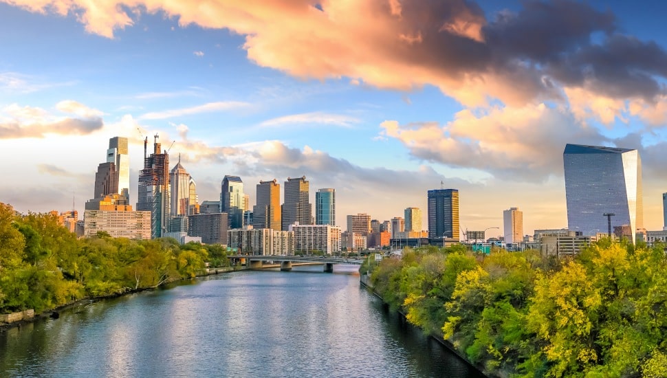 Philadelphia skyline viewed from the Schuylkill River with green trees lining the banks and dramatic clouds above Center City skyscrapers.