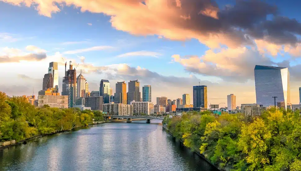 Philadelphia skyline viewed from the Schuylkill River with green trees lining the banks and dramatic clouds above Center City skyscrapers.