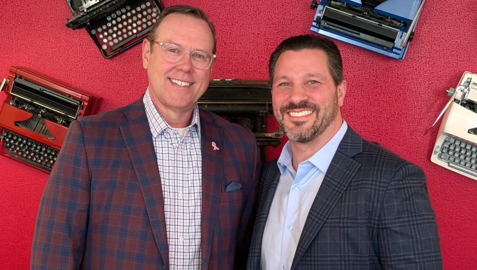 Jason Rocker and Mike Neumeier smiling together in front of a red wall decorated with vintage typewriters.