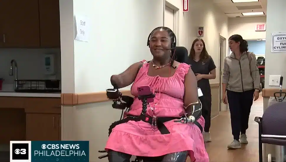 Candice Davis, a quadruple amputee and COVID-19 survivor, smiles while navigating a hallway at Jefferson Moss-Magee Rehabilitation Hospital in her power wheelchair, wearing a pink dress and an adaptive headset microphone.