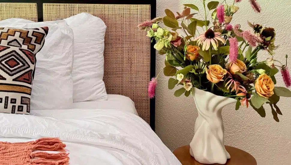 A white sculptural porcelain vase by Mount Airy ceramist Sullivan Owen holding an arrangement of orange roses, pink coneflowers, and eucalyptus, displayed on a wooden side table next to a bed.