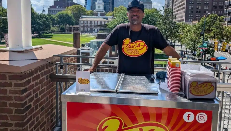 Andre Andrews stands behind a Dre's Water Ice pushcart with Independence Hall visible in the background in Philadelphia.