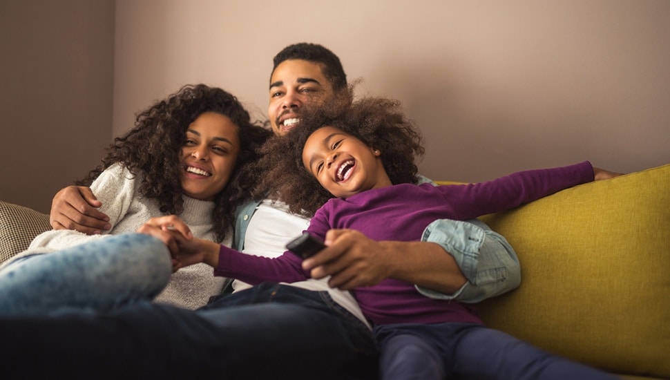 A family laughs and enjoys watching a movie on the couch.