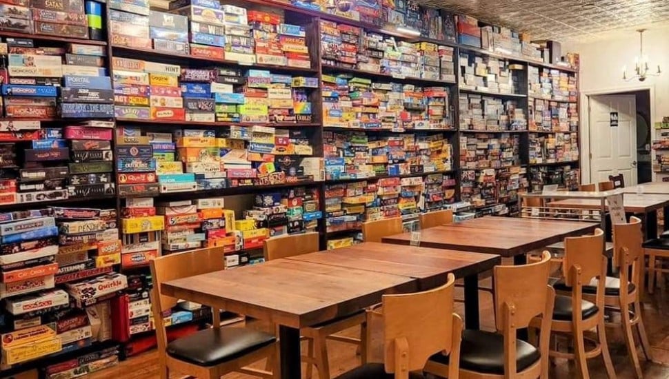 Interior of Queen & Rook Game Café in Philadelphia, showing floor-to-ceiling shelves packed with hundreds of board games alongside wooden tables and chairs.