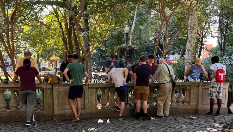 A group of people of varying ages gathered around outdoor chess tables at Rittenhouse Square in Philadelphia, surrounded by trees in summer.