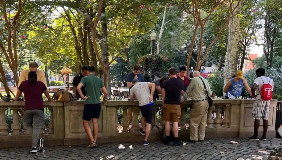 A group of people of varying ages gathered around outdoor chess tables at Rittenhouse Square in Philadelphia, surrounded by trees in summer.