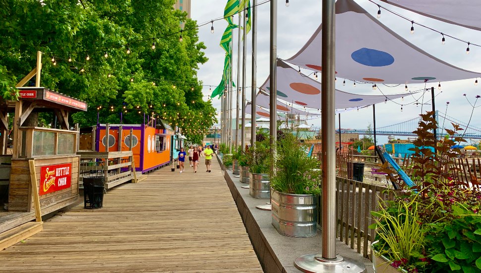 Visitors walk along the boardwalk at Spruce Street Harbor Park in Philadelphia, with colorful food stalls, string lights, and the Ben Franklin Bridge visible in the background.