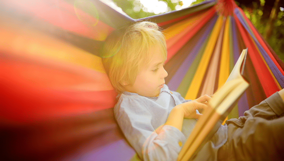 A young boy sits in a hammock reading a book.