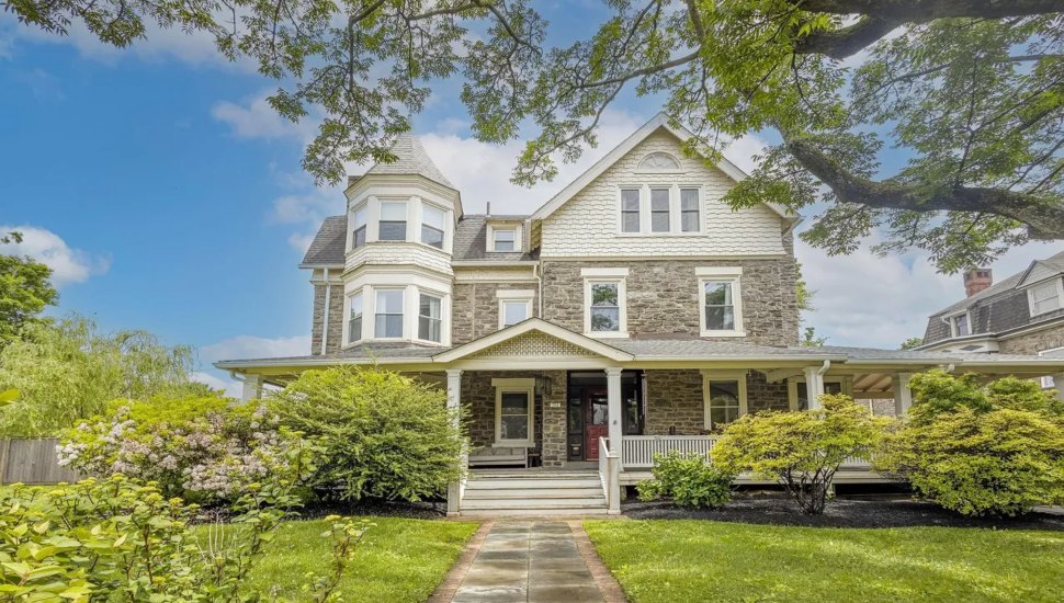 A three-story Victorian home at 106 W. Upsal St. in West Mount Airy, Philadelphia, featuring stone and shingle exterior, a wraparound porch, and lush landscaping.