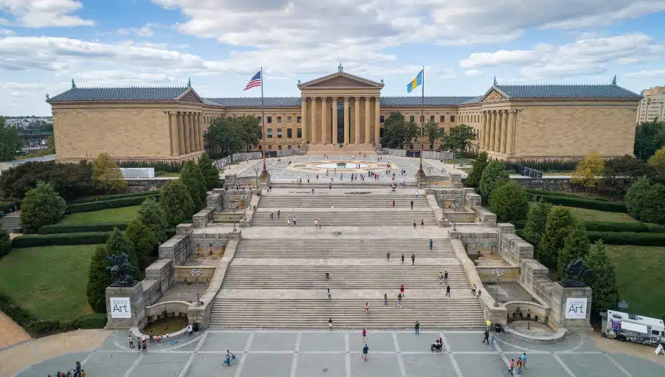 Aerial view of the Philadelphia Museum of Art and the Rocky Steps, with visitors climbing the staircase toward the museum's columned entrance flanked by American and Pennsylvania flags.