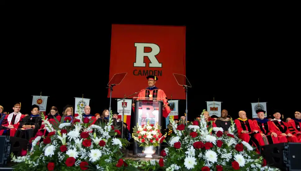 Rutgers University–Camden graduates and faculty gather on stage during the Class of 2026 commencement ceremony beneath a large Rutgers Camden banner.