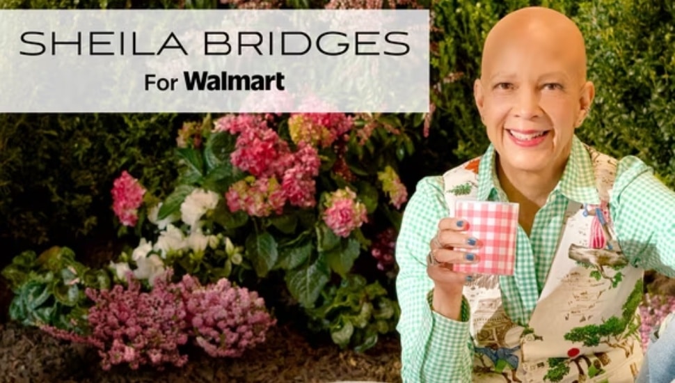 Interior designer Sheila Bridges smiles while holding a pink gingham tumbler from her Philadelphia Collection, surrounded by blooming hydrangeas in a garden setting.