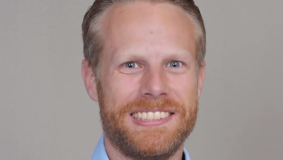 Headshot of Torben Schiffner, Ph.D., a bearded man with reddish-blond hair smiling in a light blue collared shirt against a neutral gray background.