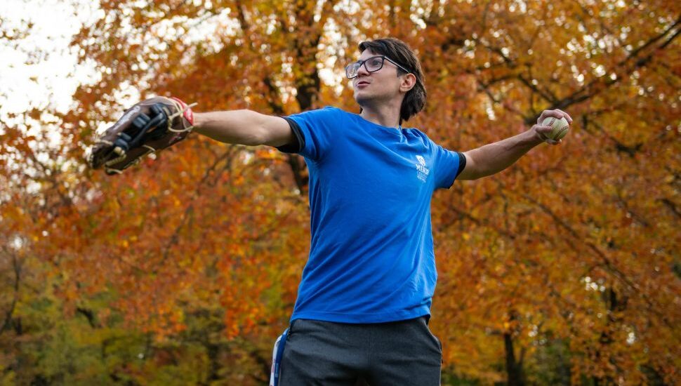 Jacob Torres Young wearing a blue Manor College shirt and baseball glove, preparing to throw a baseball in a park surrounded by vibrant autumn foliage.